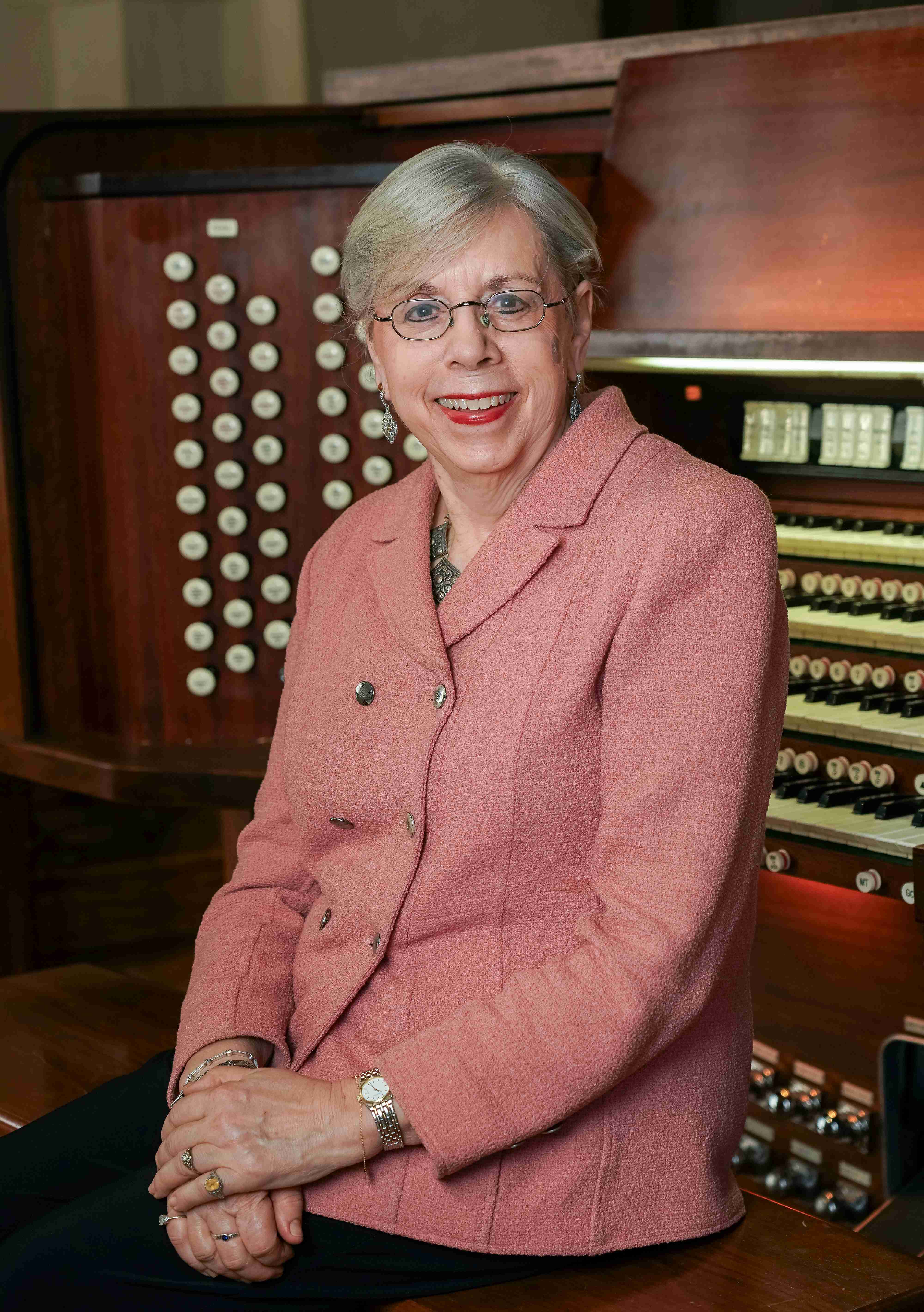 Gail Archer sits in front of an organ wearing a pink blazer. She leans on the organ and holds her hands on top of each other in front of her torso. She is smiling and looks at the camera. She wears dangling earrings, a necklace that is barely visible under the blazer, a watch and bracelets, and rings. She is a white woman who wears round rectangular wire-frame glasses. She has short grey hair that stops right above her ears. She wears eyeliner and red lipstick in the photo.